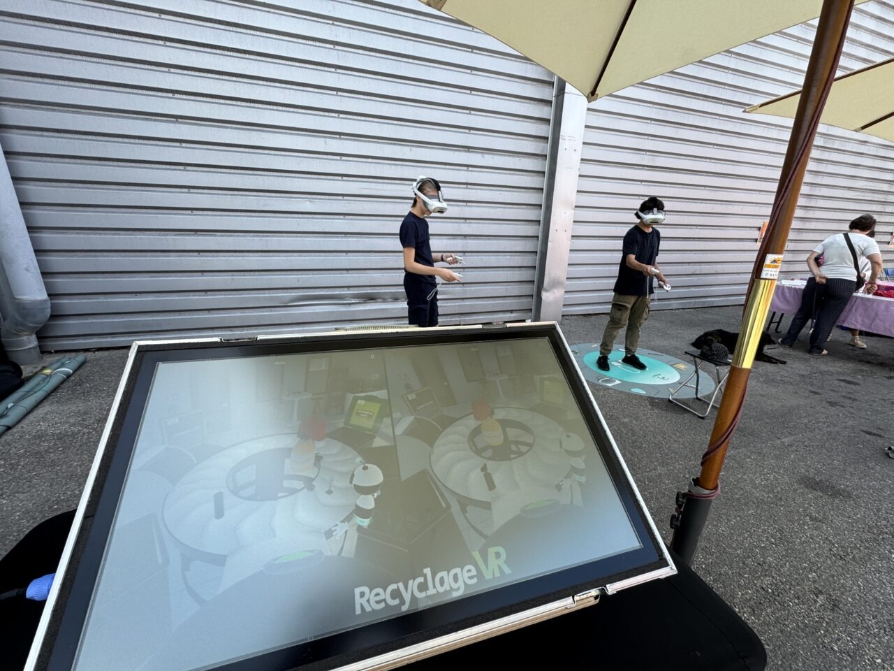 Deux personnes utilisent des casques VR sous des parasols, avec un grand écran RecyclageVR au premier plan.
