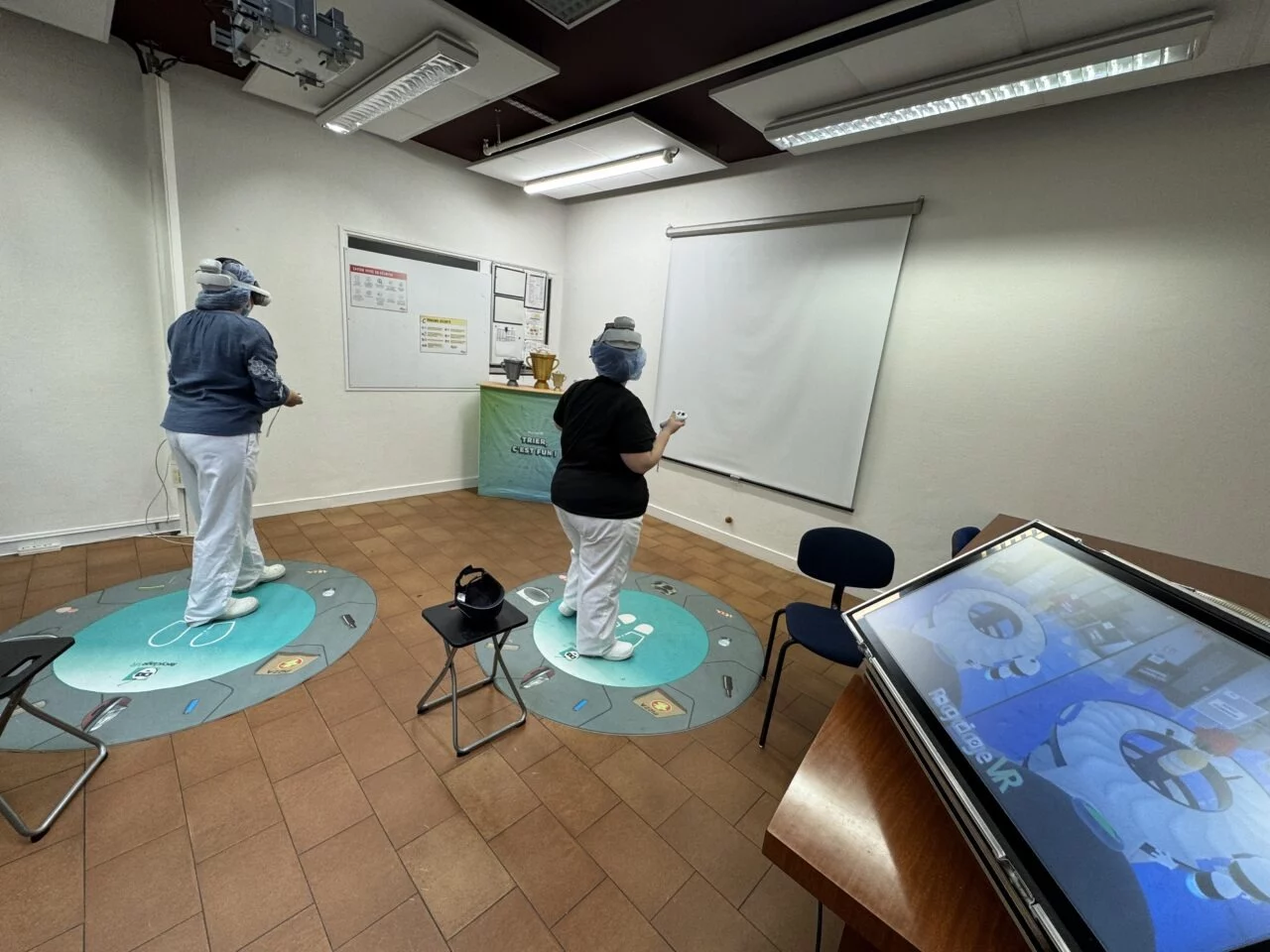 Two people using RecyclageVR headsets and controllers on mats, with a screen in the foreground
